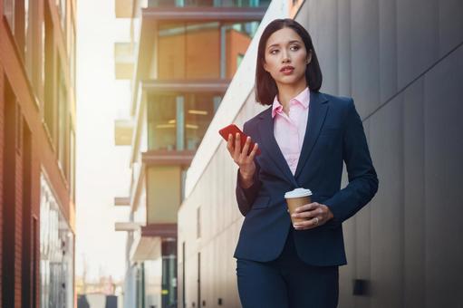 街なかでコーヒーとスマートフォンを持って立つアジア人ビジネスウーマン ビジネスウーマン,ビジネス,女性の写真素材