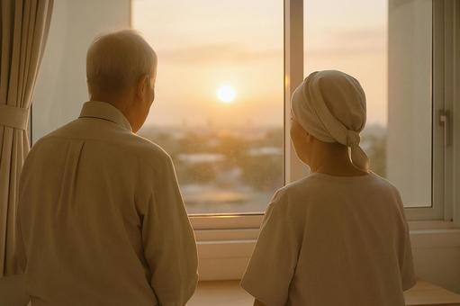病室の窓から夕陽を眺める老夫婦の写真