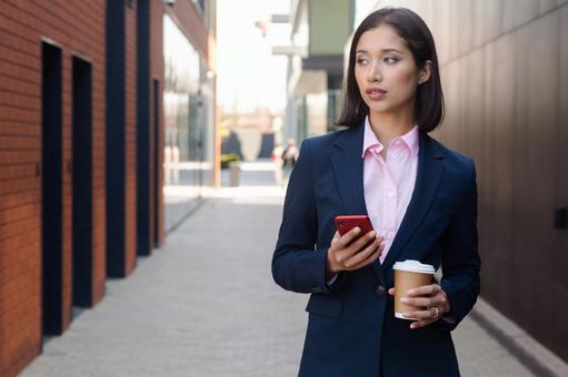 街なかでスマートフォンとコーヒーを持って立つスーツの女性 ビジネスウーマン,ビジネス,女性の写真素材