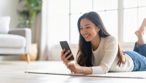 リビングで横になり携帯電話を眺める女性 リビングで横になり携帯電話を眺める女性の写真