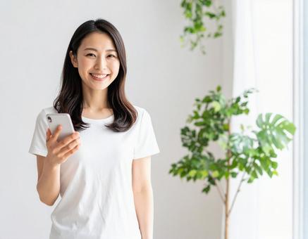 室内でスマホを持つ笑顔の白Tシャツの女性 室内でスマホを持つ笑顔の白Tシャツの女性の写真