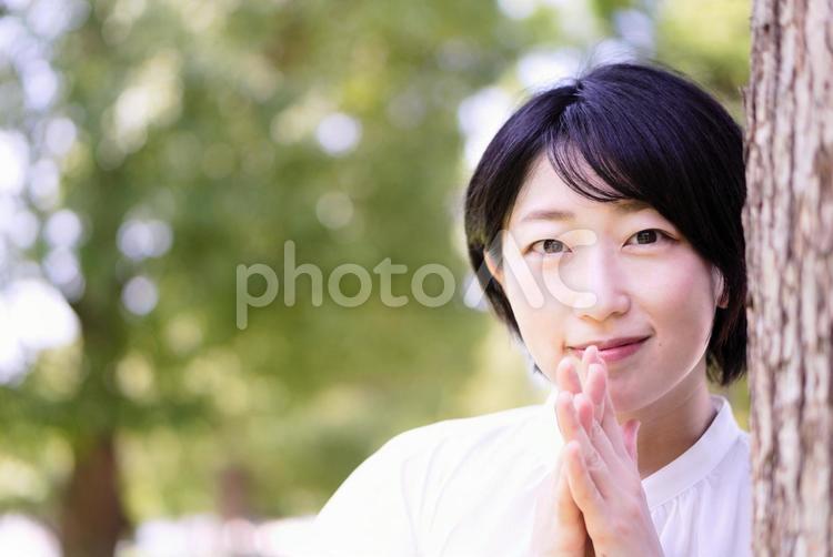 木の陰で顔の前に手を合わせる女性 女性,屋外,木の写真素材