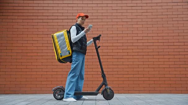電動キックボードを持ってスマートフォンを見るフードデリバリーの女性 配達,フードデリバリー,デリバリーの写真素材