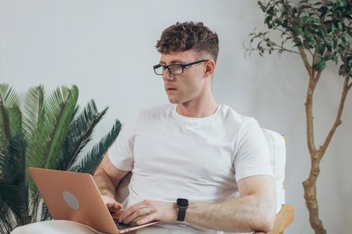 椅子に座って膝の上でノートパソコンを使う白Tシャツを着た男性 男性,外国人,欧米人の写真素材