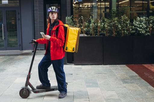 街なかでスマートフォンを手に電動キックボードに乗っているフードデリバリーの男性 配達,フードデリバリー,デリバリーの写真素材