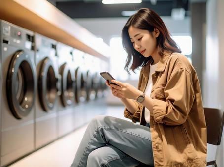 コインランドリーでスマホを見る若い女性 コインランドリーでスマホを見る若い女性の写真