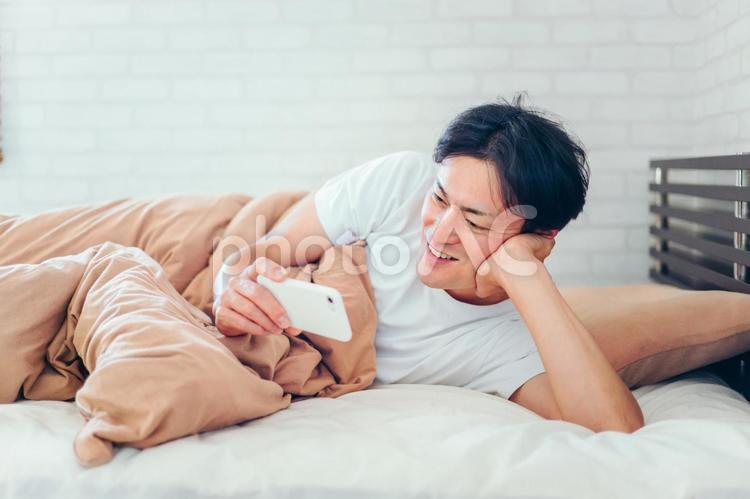 ベッドで寝ながらスマホを見る男性（笑顔） ベッド,寝る前スマホ,アプリの写真素材
