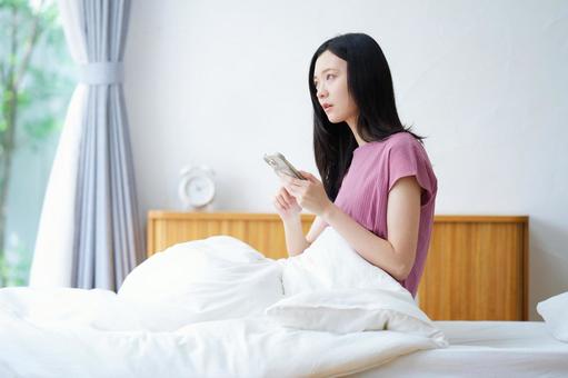 ベッドでスマホを持ち外を見つめる女性 女性,スマートフォン,持つの写真素材