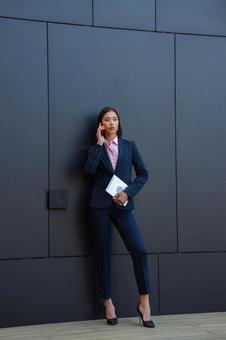 オフィスビルの黒い壁の前で電話しているパンツスーツのアジア人女性 ビジネスウーマン,ビジネス,女性の写真素材