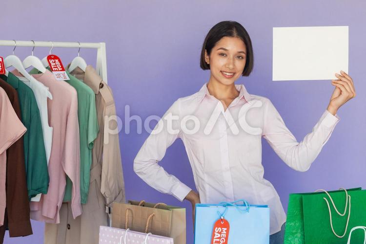 セールのタグの付いたハンガーに掛かった洋服の前で白い紙を見せる女性 セール,ショップ,女性の写真素材
