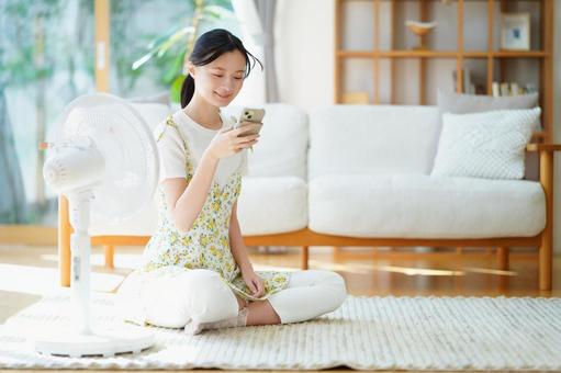 扇風機の前に座りスマホを見る笑顔女性 女性,扇風機,スマートフォンの写真素材