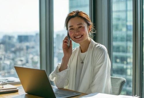 パソコンをしながら笑顔で電話する女性 パソコンをしながら笑顔で電話する女性の写真
