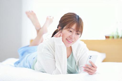 ベッドに寝転び笑顔でスマホを見る女性 女性,スマートフォン,持つの写真素材