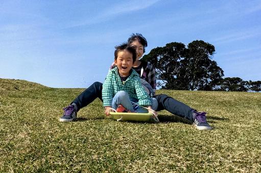 <素材>公園の斜面で草スキーをする親子 <素材>公園の斜面で草スキーをする親子 家族,子供,女性の写真素材