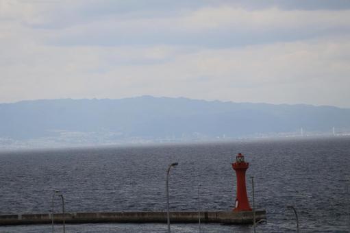 赤い灯台と突堤と海と空と霞む街と山の写真