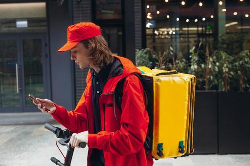 街なかでスマートフォンを見ている電動キックボードに乗るフードデリバリーの男性 配達,フードデリバリー,デリバリーの写真素材