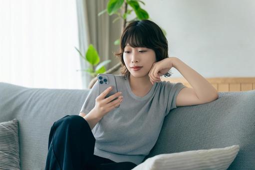 ソファに座ってスマホを使う笑顔の女性 ソファに座ってスマホを使う笑顔の女性の写真