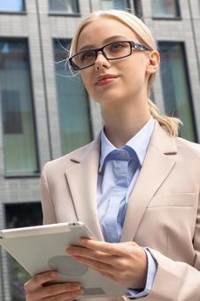 オフィスビルの外でタブレットを持って立つメガネを掛けた女性 ビジネスウーマン,ビジネス,女性の写真素材