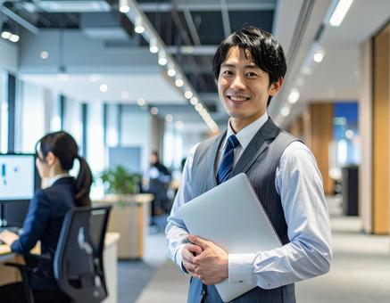 オフィスでPCを持って微笑む男性社員 オフィスでPCを持って微笑む男性社員の写真