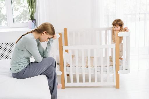 悩む女性とベッドの柵に掴まり立つ子供 悩む女性とベッドの柵に掴まり立つ子供 育児疲れ,疲れた,座るの写真素材