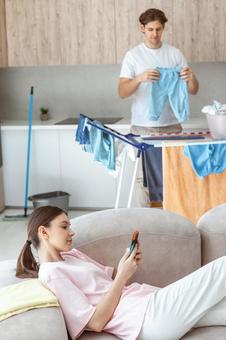 横になって携帯を見る女性と洗濯をする男性 横になって携帯を見る女性と洗濯をする男性 夫婦,女性,人物の写真素材