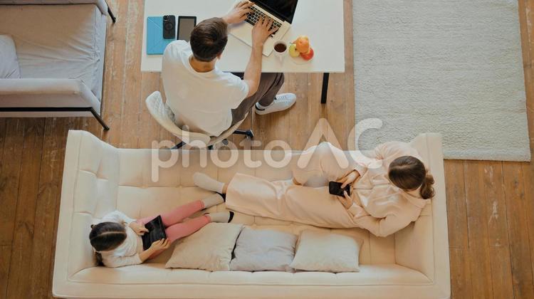 男性が仕事をしていてソファーで携帯をする女性とタブレットをしてくつろぐ女の子 家族,男性,女の子の写真素材