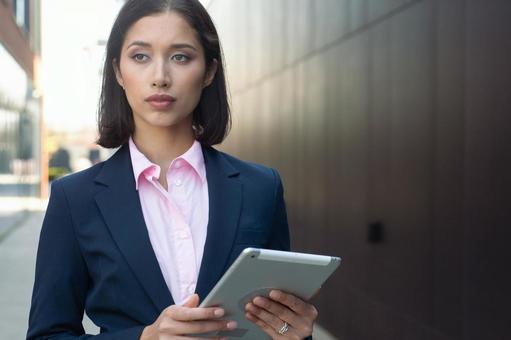 街なかでタブレットを持って立つスーツのアジア人女性 ビジネスウーマン,ビジネス,女性の写真素材