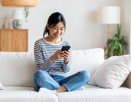 ソファに座ってスマホを見て笑う女性 ソファに座ってスマホを見て笑う女性の写真