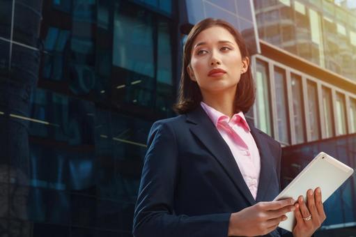 オフィスビルの外でスーツのアジア人女性がタブレットを持って立っている ビジネスウーマン,ビジネス,女性の写真素材