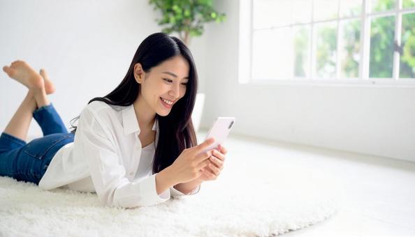 リビングで横になりスマホを操作する女性 リビングで横になりスマホを操作する女性の写真