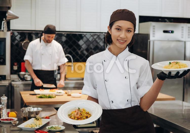 出来上がった料理を見せる女性シェフと調理中の男性シェフ キュイジーヌ,キッチン,厨房の写真素材