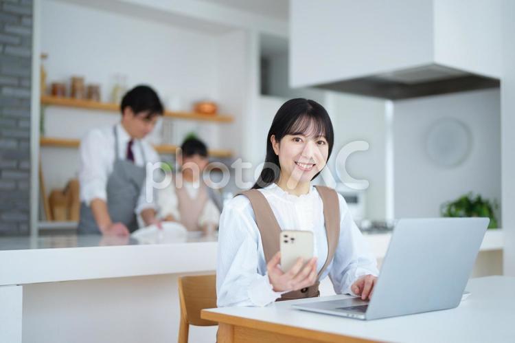 携帯を持ってパソコンで仕事する女性と後ろで皿洗いする親子 家族,女の子,女性の写真素材