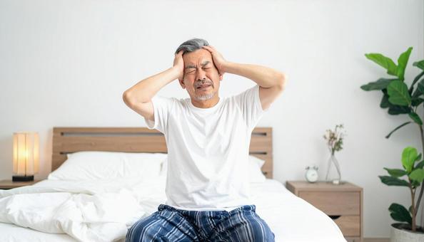 寝起きで座って頭を抱えるパジャマ姿の男性 寝起きで座って頭を抱えるパジャマ姿の男性の写真