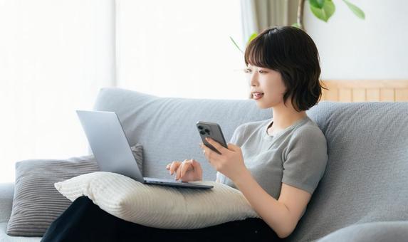 ソファに座ってスマホとパソコンを使う女性 女性,ソファ,パソコンの写真素材