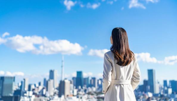 青空の下、遠くの景色を眺める女性の後ろ姿の写真