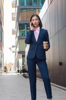 街なかでコーヒー片手に電話するパンツスーツの女性 ビジネスウーマン,ビジネス,女性の写真素材