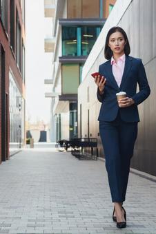 街なかでコーヒーとスマートフォンを持って歩くパンツスーツの女性 ビジネスウーマン,ビジネス,女性の写真素材