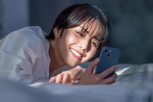ベッドに寝てスマートフォンを操作する女性 ベッドに寝てスマートフォンを操作する女性 スマホ,スマートフォン,検索の写真素材