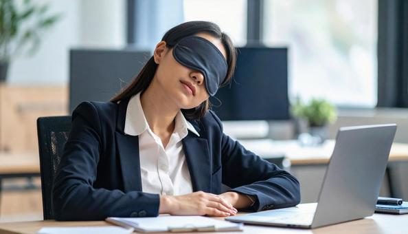 アイマスクをしてデスクで仮眠する女性の写真