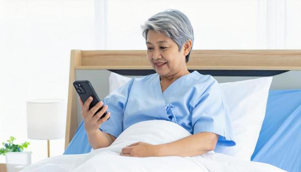 ベッドに座り携帯を見る日本人シニア女性の写真