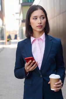 街なかでコーヒーとスマートフォンを持って立つスーツのアジア人女性 ビジネスウーマン,ビジネス,女性の写真素材