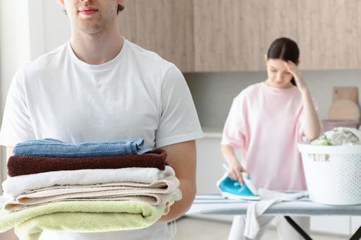 タオルを持った男性と後ろでアイロンする女性 タオルを持った男性と後ろでアイロンする女性 夫婦,女性,人物の写真素材