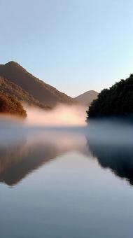 静寂に包まれた湖、夜明けの光に染まる朝霧の写真