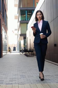 街なかでコーヒーとスマートフォンを持って歩いているパンツスーツの女性 ビジネスウーマン,ビジネス,女性の写真素材