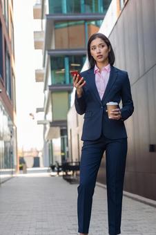 街なかでコーヒーとスマートフォンを持って立つビジネスウーマン ビジネスウーマン,ビジネス,女性の写真素材