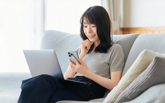 ソファに座ってスマホとパソコンを使う女性 ソファに座ってスマホとパソコンを使う女性の写真