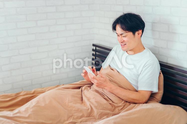 ベッドで寝ながらスマホを見る男性（笑顔） ベッド,寝る前スマホ,アプリの写真素材
