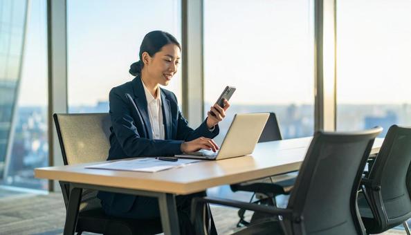 スマホを持ちながらPC作業をする女性 スマホを持ちながらPC作業をする女性の写真