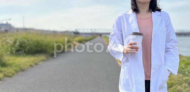 コーヒー片手に河原を歩く女医。ほっと一息 白衣,先生,医師の写真素材