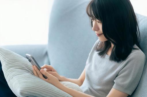 ソファに座ってスマホを使う笑顔の女性 ソファに座ってスマホを使う笑顔の女性 女性,スマホ,スマートフォンの写真素材